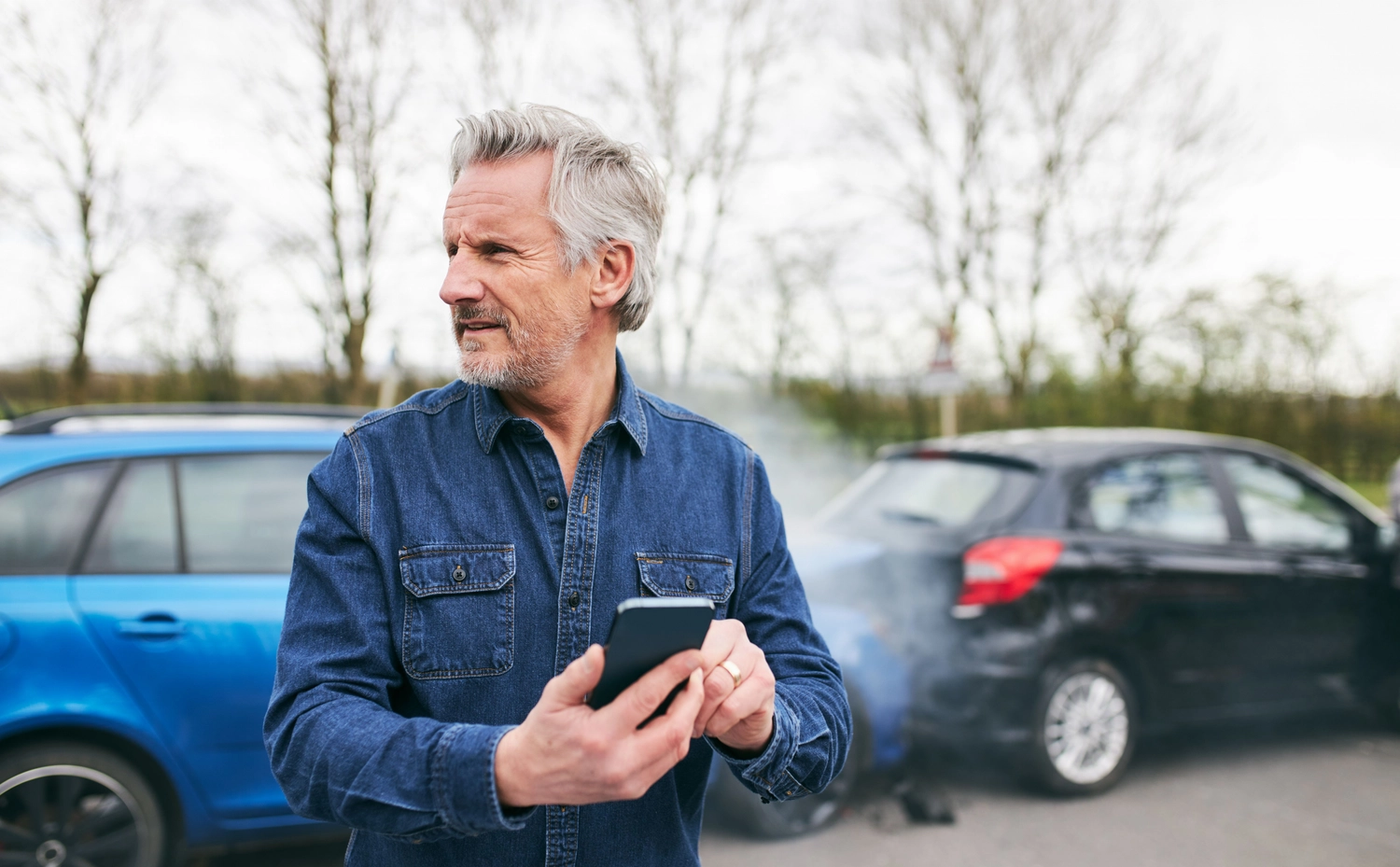 SVH Sachverständigenbüro – Kfz-Experte – Ein Mann hält ein Smartphone vor zwei kollidierten Fahrzeugen, aus dem hinteren Fahrzeug steigt Rauch auf. Nach einem Unfall haben Ihre Sicherheit und der rechtliche Schutz oberste Priorität.